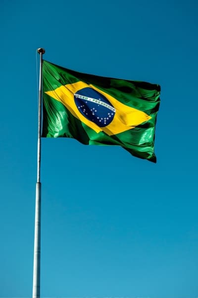 Imagem da bandeira do brasil hasteada contra um céu azul, ao fundo.